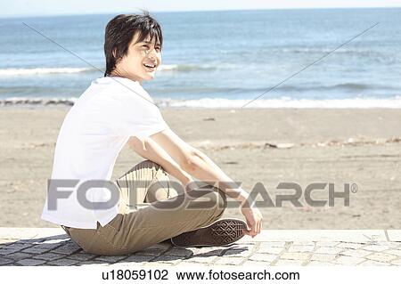 Stock Image - Japanese man sitting on the beach. Fotosearch