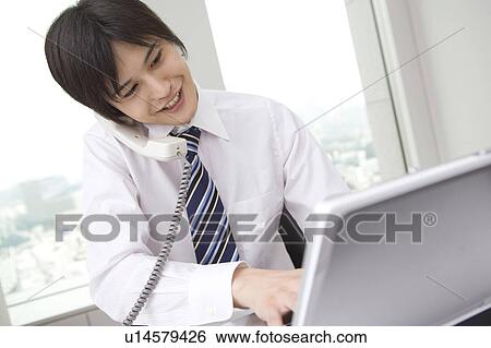 Stock Photograph - Office worker talking on phone while operating laptop computer. Fotosearch