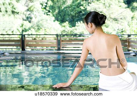 Stock Photo - Young woman with towel sitting by pool, rear view. Fotosearch