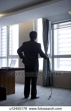 Stock Photo - Businessman holding golf club looking through window in office. Fotosearch
