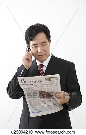 Stock Image - Businessman on the phone looking at newspaper in studio shot. Fotosearch