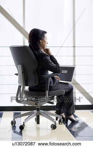 Businesswoman sitting on chair with laptop on lap by window View Large Photo Image Stock Photo - Businesswoman sitting on chair with laptop on lap by window. Fotosearch
