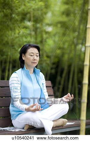 Stock Image - Senior woman sitting on bench on lotus position in park. Fotosearch