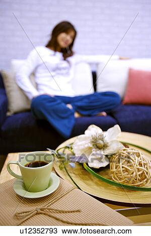 Stock Image - Young woman sitting on sofa, a cup of coffee in the foreground. Fotosearch