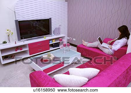 Stock Image - Young woman sitting on sofa using laptop in living room. Fotosearch