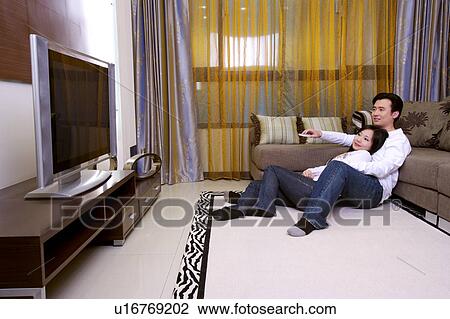 Stock Image - Couple sitting on floor leaning on sofa watching TV. Fotosearch
