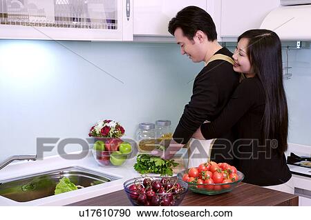 Stock Image - Woman hugging man as he prepares food in domestic kitchen. Fotosearch