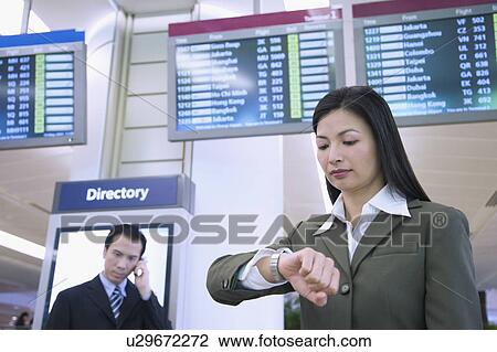 Stock Image - Low angle view of a businesswoman looking at her wristwatch with a businessman talking on a mobile phone behind her at an airport. Fotosearch