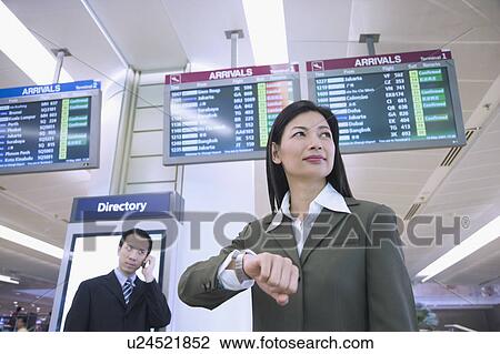 Stock Image - Low angle view of a businesswoman with a businessman talking on a mobile phone behind her at an airport. Fotosearch