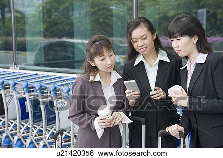 Stock Photograph - Three businesswomen looking at a personal data assistant at an airport lounge. Fotosearch