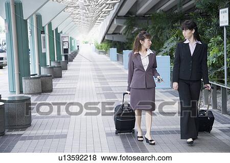 Two businesswomen walking on a walkway with their luggage View Large Photo Image Stock Photo - Two businesswomen walking on a walkway with their luggage. Fotosearch
