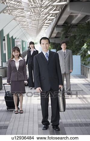 Picture - Businessman walking and holding his suitcase with three business executives in the background. Fotosearch