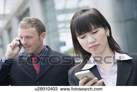 Close-up of a businesswoman using a personal data assistant with a businessman talking on a mobile phone behind her View Large Photo Image Stock Image - Close-up of a businesswoman using a personal data assistant with a businessman talking on a mobile phone behind her. Fotosearch