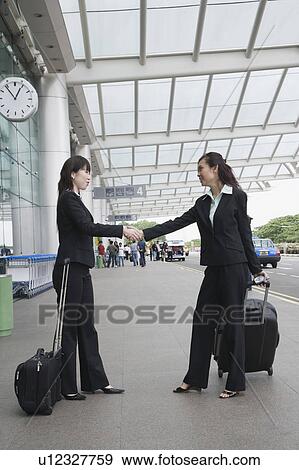 Stock Photo - Side profile of two businesswomen shaking hands outside an airport. Fotosearch