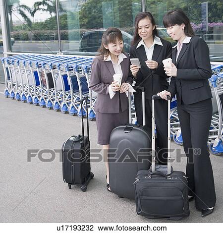 Stock Image - Three businesswomen looking at a personal data assistant at an airport lounge. Fotosearch