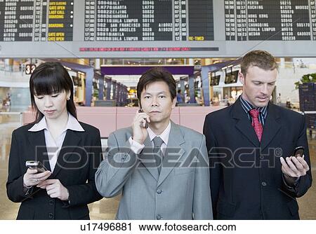 Stock Image - Two businessmen and a businesswoman holding mobile phones and standing at an airport. Fotosearch