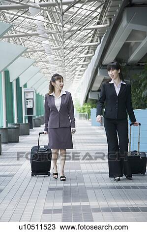 Stock Image - Two businesswomen walking on a walkway with their luggage. Fotosearch