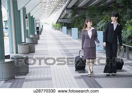 Two businesswomen walking on a walkway with their luggage View Large Photo Image Stock Image - Two businesswomen walking on a walkway with their luggage. Fotosearch