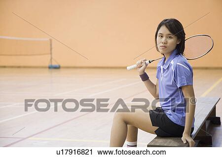 Stock Image - Portrait of a young woman sitting on a bench and holding a badminton racket. Fotosearch