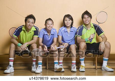 Stock Photo - Portrait of two young women and two young men sitting side by side on a bench and holding badminton rackets. Fotosearch