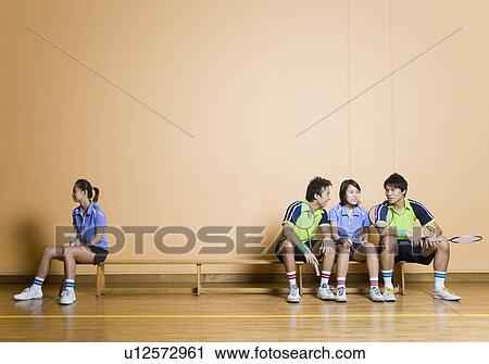 Stock Image - Two young men and a young woman sitting side by side on a bench with another young woman sitting at other corner. Fotosearch
