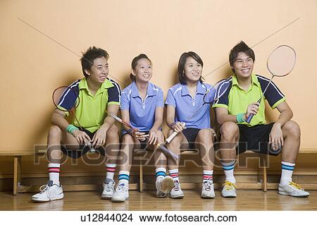 Picture - Two young women and two young men sitting side by side on a bench and holding badminton rackets. Fotosearch