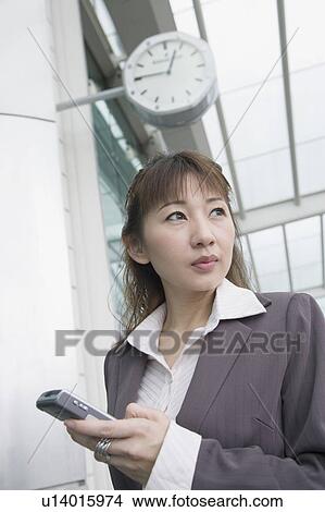 Picture - Low angle view of a businesswoman holding a mobile phone and waiting at an airport lounge. Fotosearch