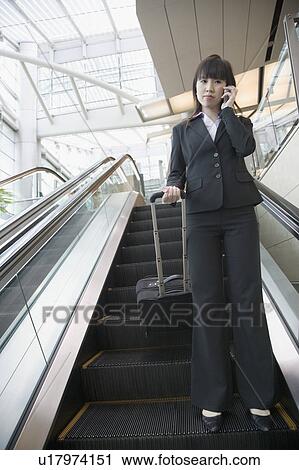 Stock Image - Low angle view of a businesswoman standing on an escalator and talking on a mobile phone. Fotosearch