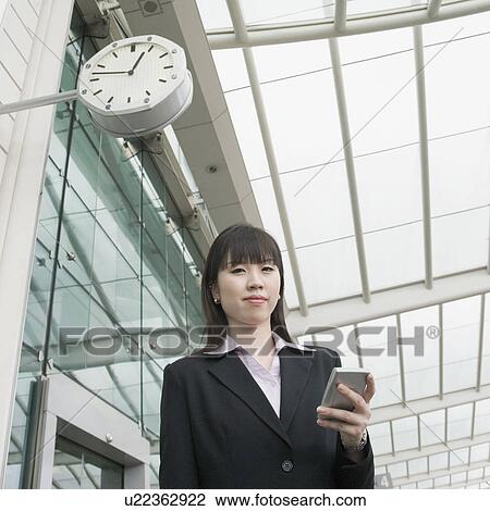 Stock Image - Low angle view of a businesswoman holding a personal data assistant and waiting at an airport lounge. Fotosearch