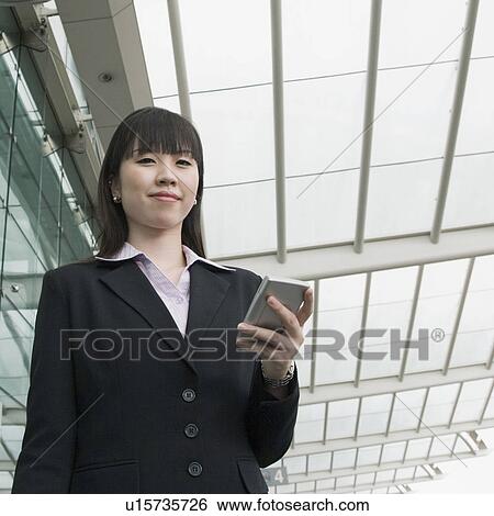 Stock Photograph - Low angle view of a businesswoman holding a personal data assistant and waiting at an airport lounge. Fotosearch