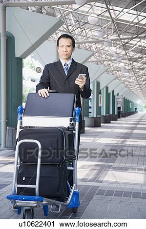 Stock Image - Businessman using a palmtop and pushing a luggage cart. Fotosearch