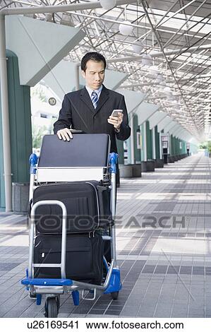 Stock Image - Businessman using a palmtop and pushing a luggage cart. Fotosearch