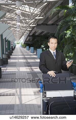 Stock Photo - Businessman using a palmtop and pushing a luggage cart. Fotosearch