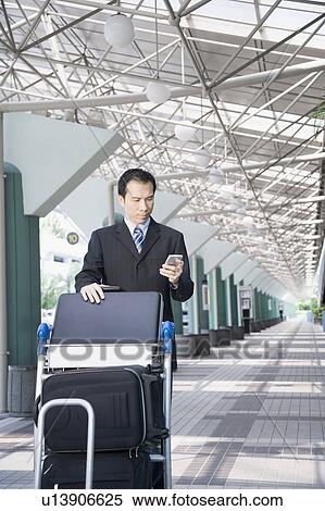 Stock Photography - Businessman using a palmtop and pushing a luggage cart. Fotosearch