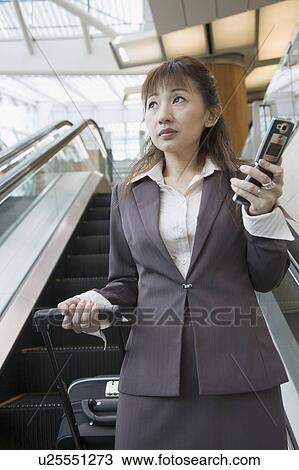 Stock Image - Low angle view of a businesswoman holding a mobile phone and a suitcase. Fotosearch
