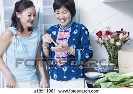 Stock Image - Senior woman preparing food with her granddaughter standing behind her and smiling. Fotosearch