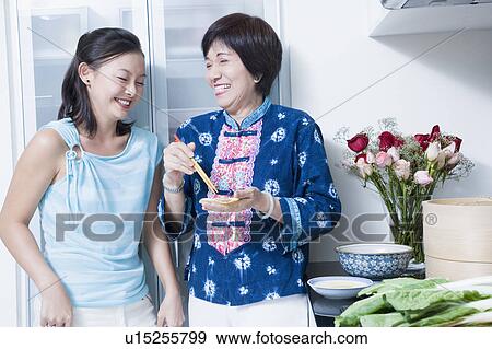 Stock Photo - Senior woman preparing food with her granddaughter standing beside her and smiling. Fotosearch