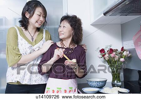 Stock Photograph - Senior woman preparing food with her granddaughter standing behind her and smiling. Fotosearch