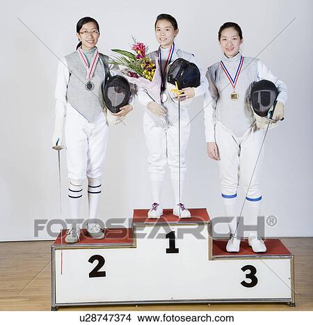 Picture - Portrait of three young women in fencing costumes standing on a winner's podium. Fotosearch