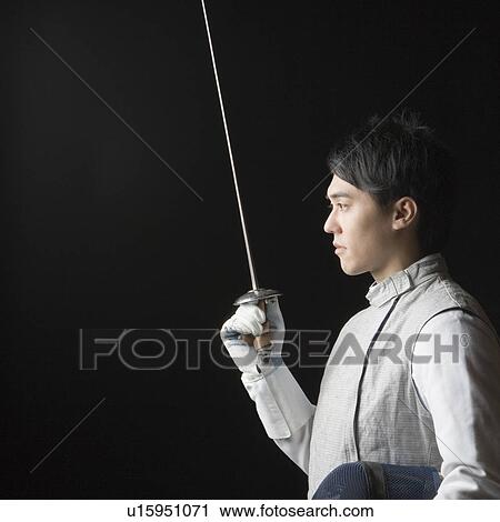 Stock Image - Side profile of a male fencer holding a fencing foil and a fencing mask. Fotosearch