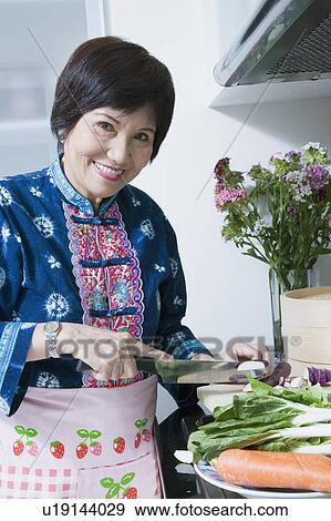 Stock Photo - Portrait of a senior woman cutting vegetables and smiling in a kitchen. Fotosearch