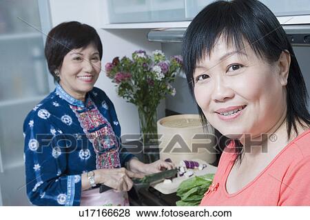 Stock Photo - Portrait of a mature woman smiling with her mother cutting vegetables in a kitchen. Fotosearch