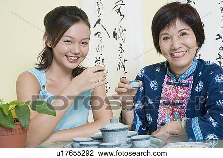 Stock Image - Portrait of a senior woman sitting with her granddaughter at a table and holding tea cups. Fotosearch