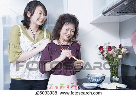Stock Photo - Senior woman preparing food with her granddaughter standing behind her and smiling. Fotosearch