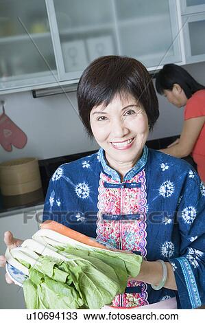 Stock Image - Portrait of a senior woman holding vegetables and smiling in a domestic kitchen. Fotosearch