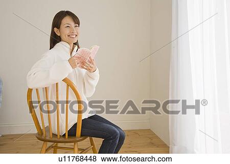 Stock Photo - Young woman opening book on chair. Fotosearch
