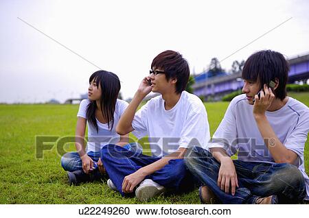 Stock Image - Three teenagers sitting on the lawn, using mobile phone and looking away, Teenager. Fotosearch