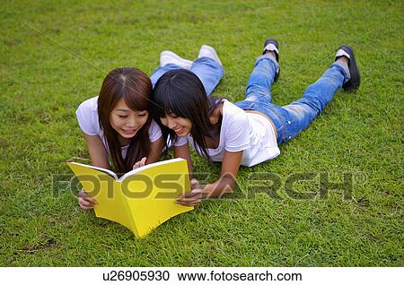 Stock Image - Two girls lying on the lawn and reading a book together, Teenager. Fotosearch