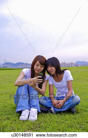 Stock Image - Two girls sitting on the lawn and looking at the mobile phone, Teenager. Fotosearch