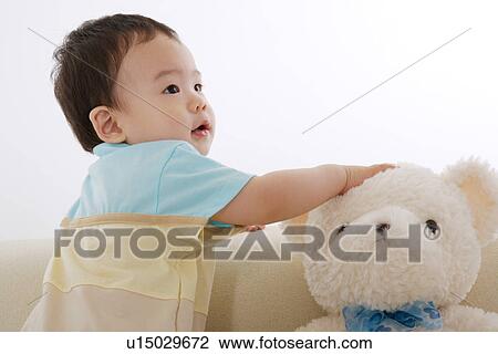 Stock Image - Baby boy on sofa touching teddy bear looking away. Fotosearch